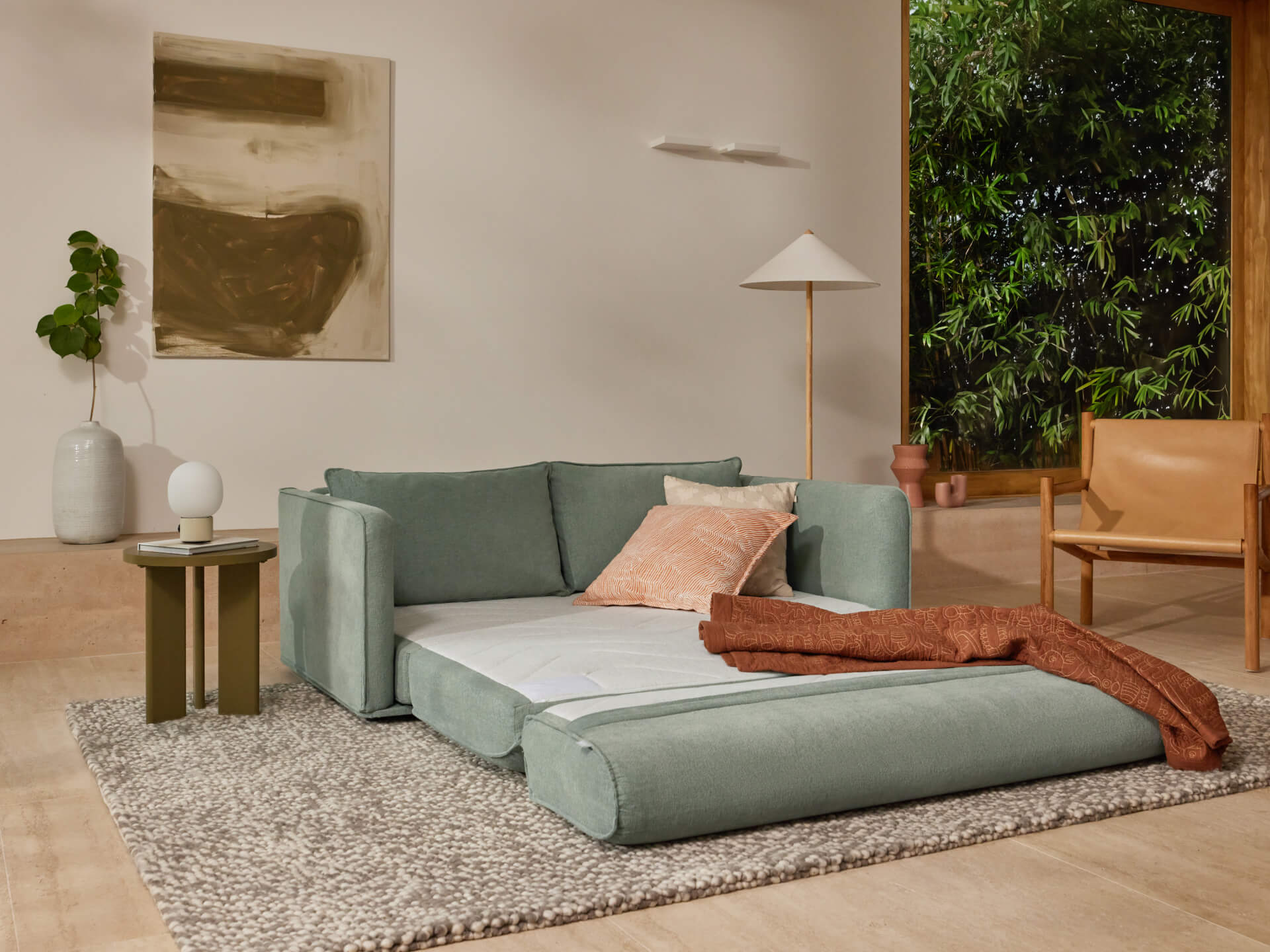 A light green sofa bed extended on a textured grey rug, with a pink pillow and brown throw. Beige wall, art, lamp, and greenery in the background.