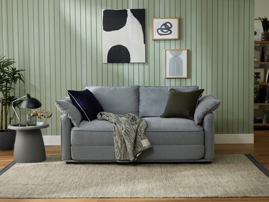 Grey sofa with assorted cushions and a patterned throw. Set against a green panelled wall with abstract art. Grey side table and lamp nearby.