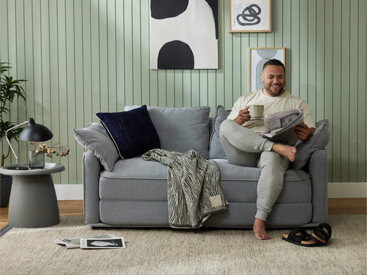 Man relaxing on light grey sofa with cushions, reading a newspaper. Green panelled wall, abstract art, lamp, and side table in the background.