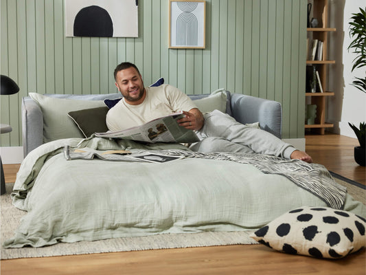 Man reading on a light grey modular sofa bed with sage green bedding in a cosy room with green panelled walls and modern art.