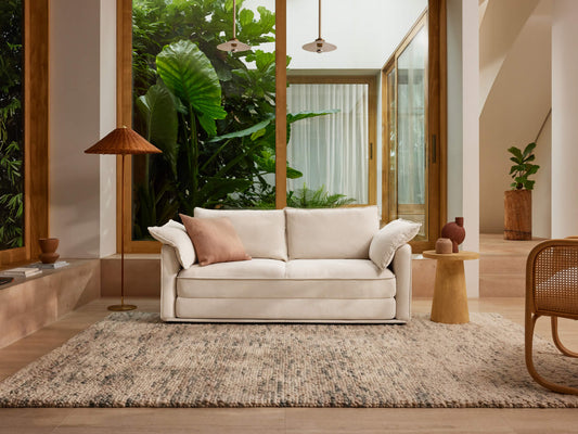 Modern living room with a white sofa, pink cushion, large green plants outside, wooden accents, tan chair, and woven rug.