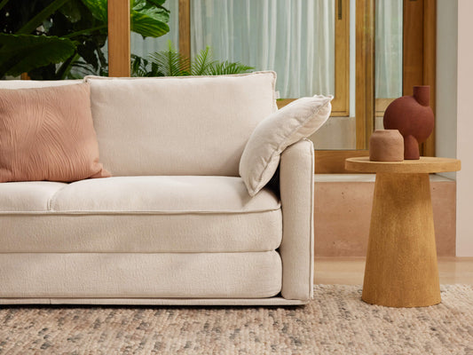 Cream sofa with a pink cushion, beside a wooden side table holding two brown vases. There are plants and a window in the background.