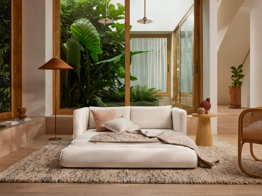 Modern living room with white sofa bed, beige and pink cushions, a brown woven rug, wooden side table, floor lamp, and large glass windows.