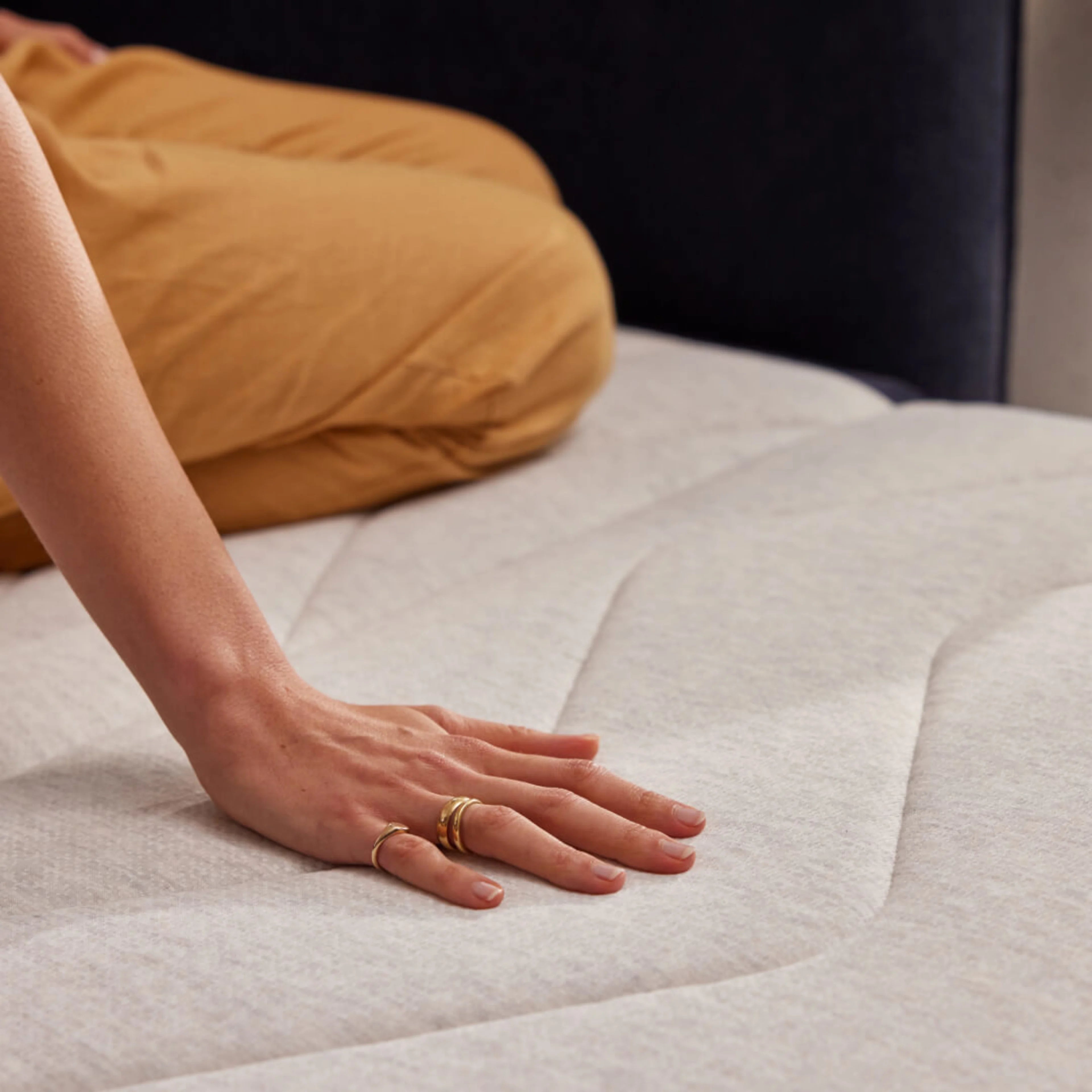 Hand pressing down on a light grey mattress, showing texture and comfort, with a person in brown trousers seated in the background.