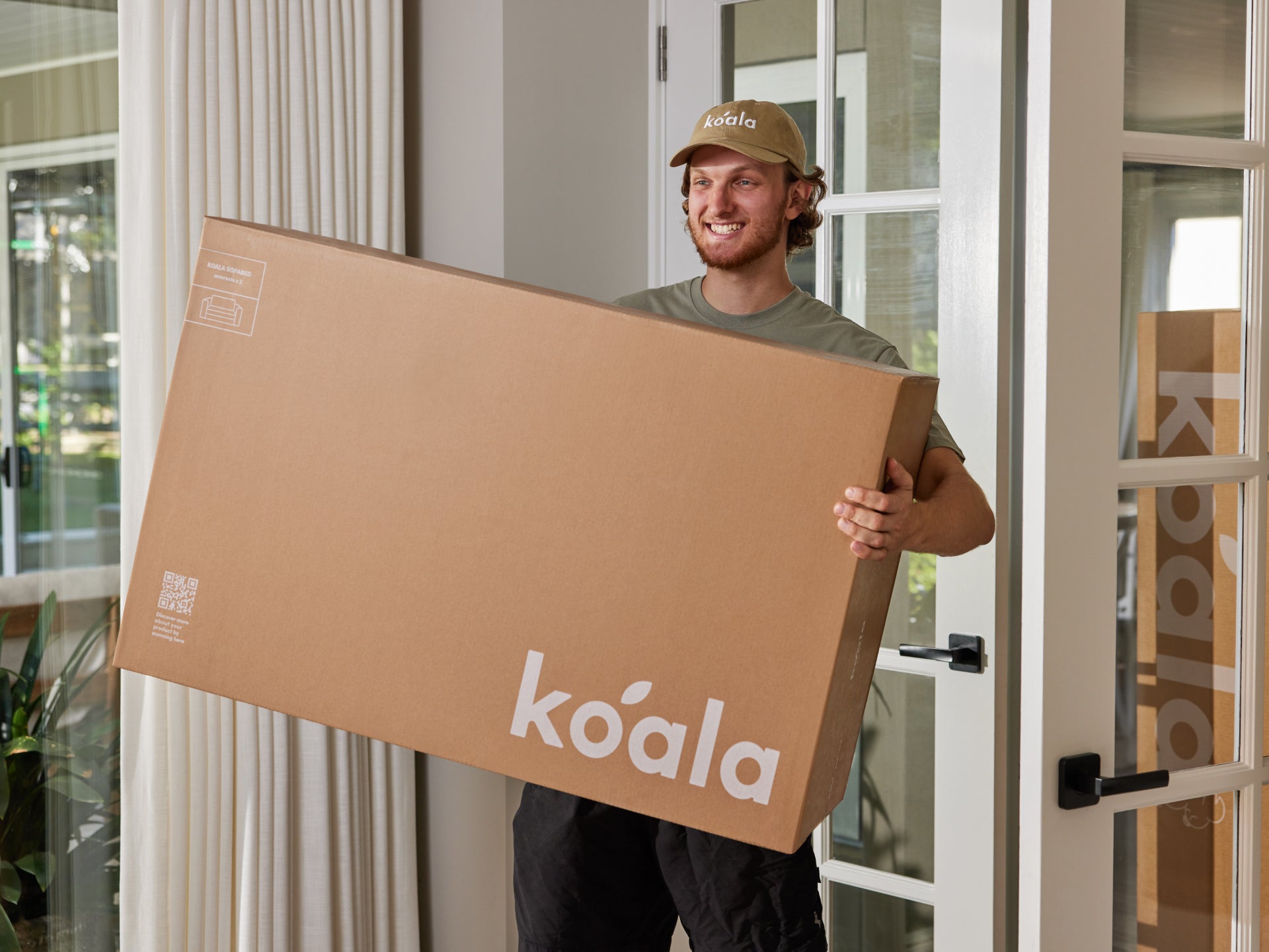 Smiling person carries a large brown box with "koala" logo in a bright room with glass doors and white curtains.