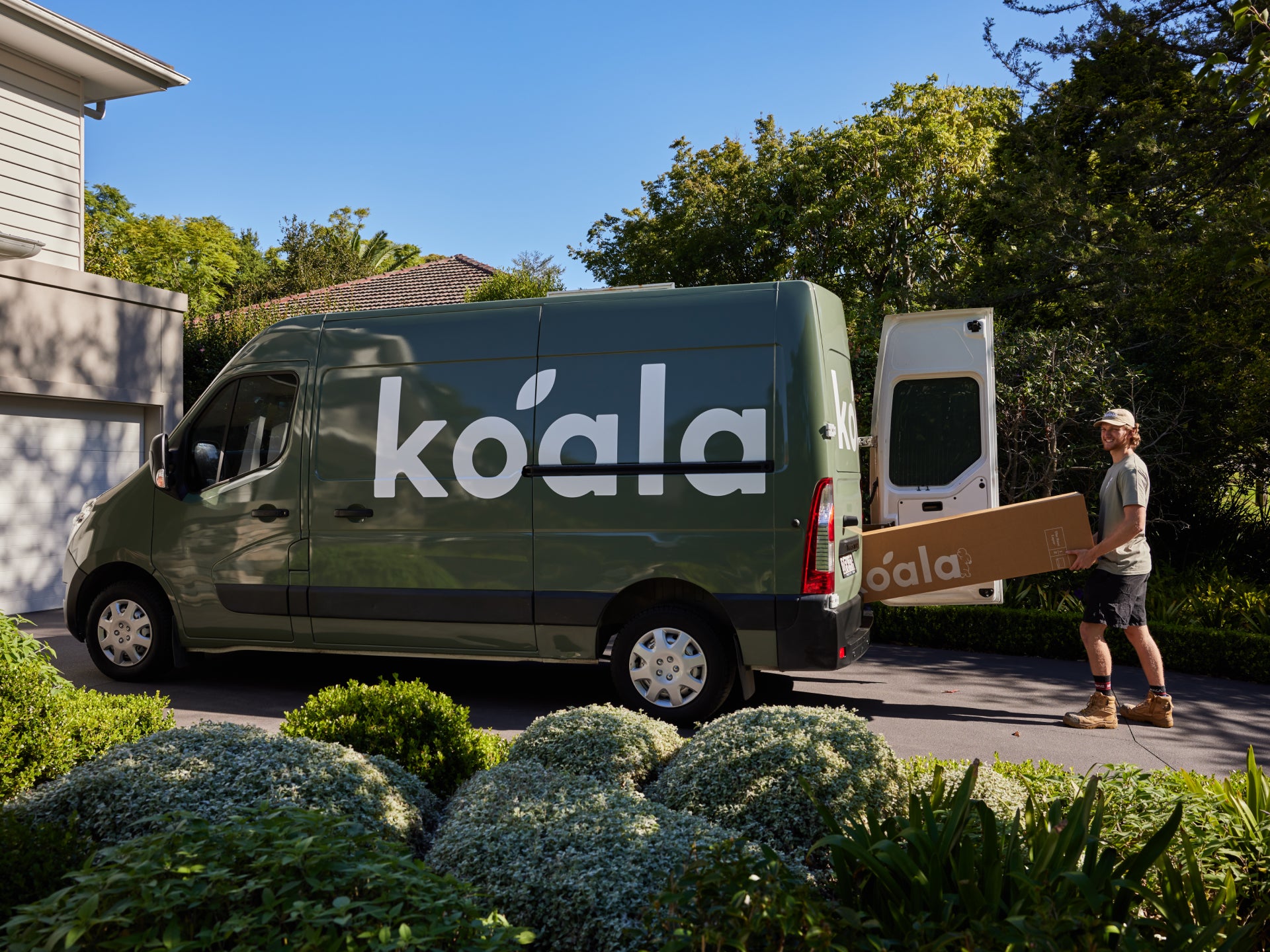 Green delivery van with "koala" branding parked in a driveway. A person unloads a large brown box from the van. Lush greenery surrounds the scene.