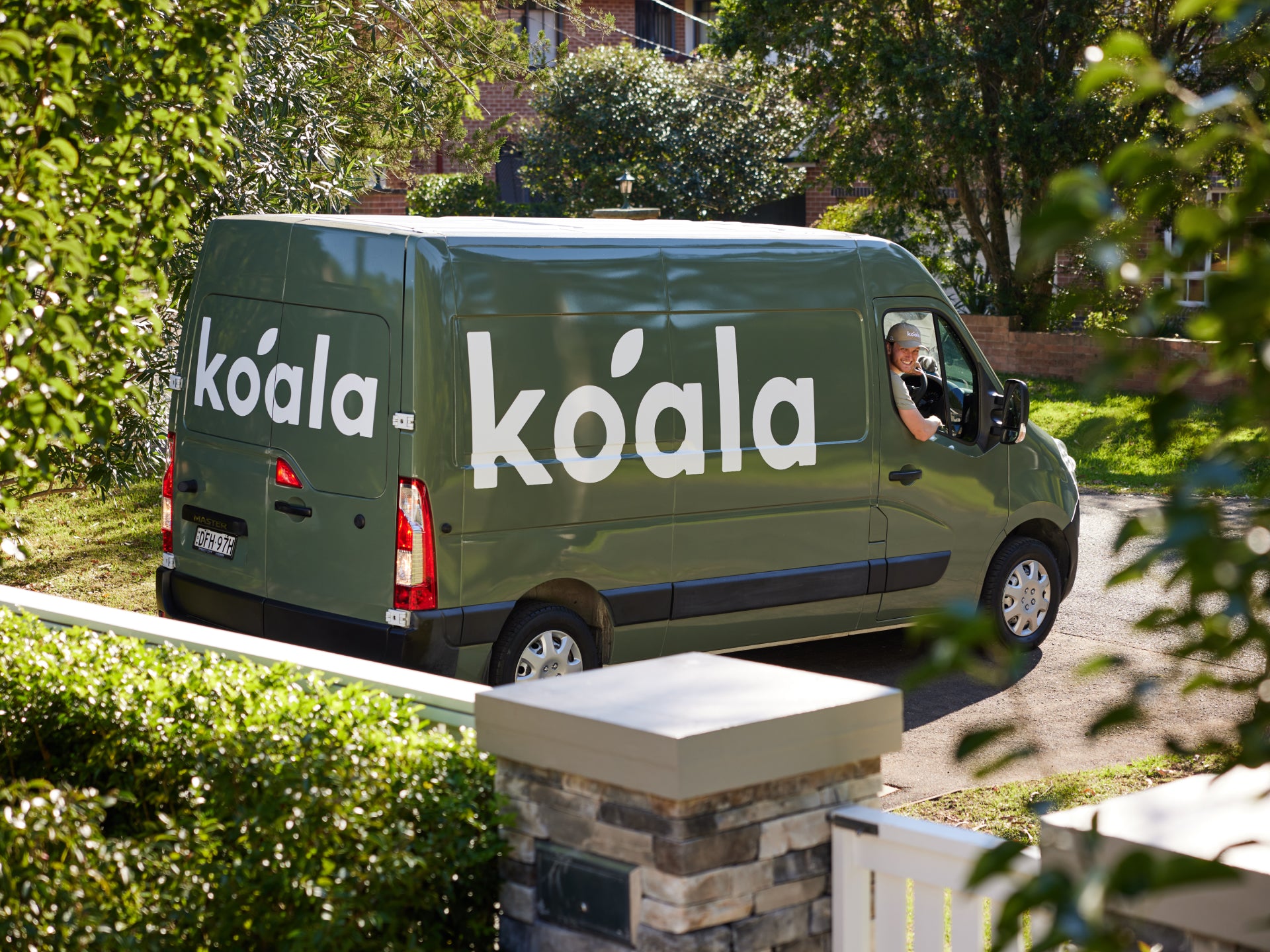 Green delivery van with "Koala" written in white, parked in a leafy suburban area, surrounded by trees and a brick fence.