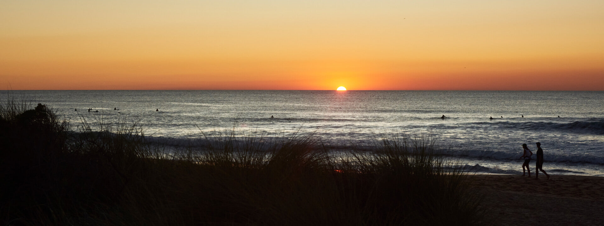 Sunset over the ocean with silhouettes of plants in the foreground and people walking along the beach under an orange and yellow sky.