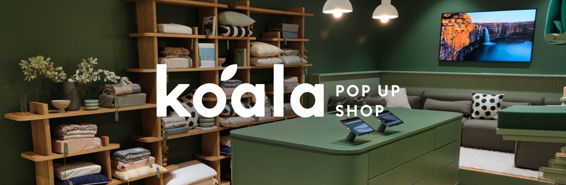 Green koala pop-up shop with wooden shelves, neatly stacked textiles, a green counter, and a sofa. Wall-mounted TV shows a nature scene.