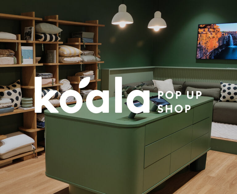 Green-themed Koala pop-up shop with wooden shelves displaying pillows and bedding, a green counter, pendant lights, and a wall-mounted TV.