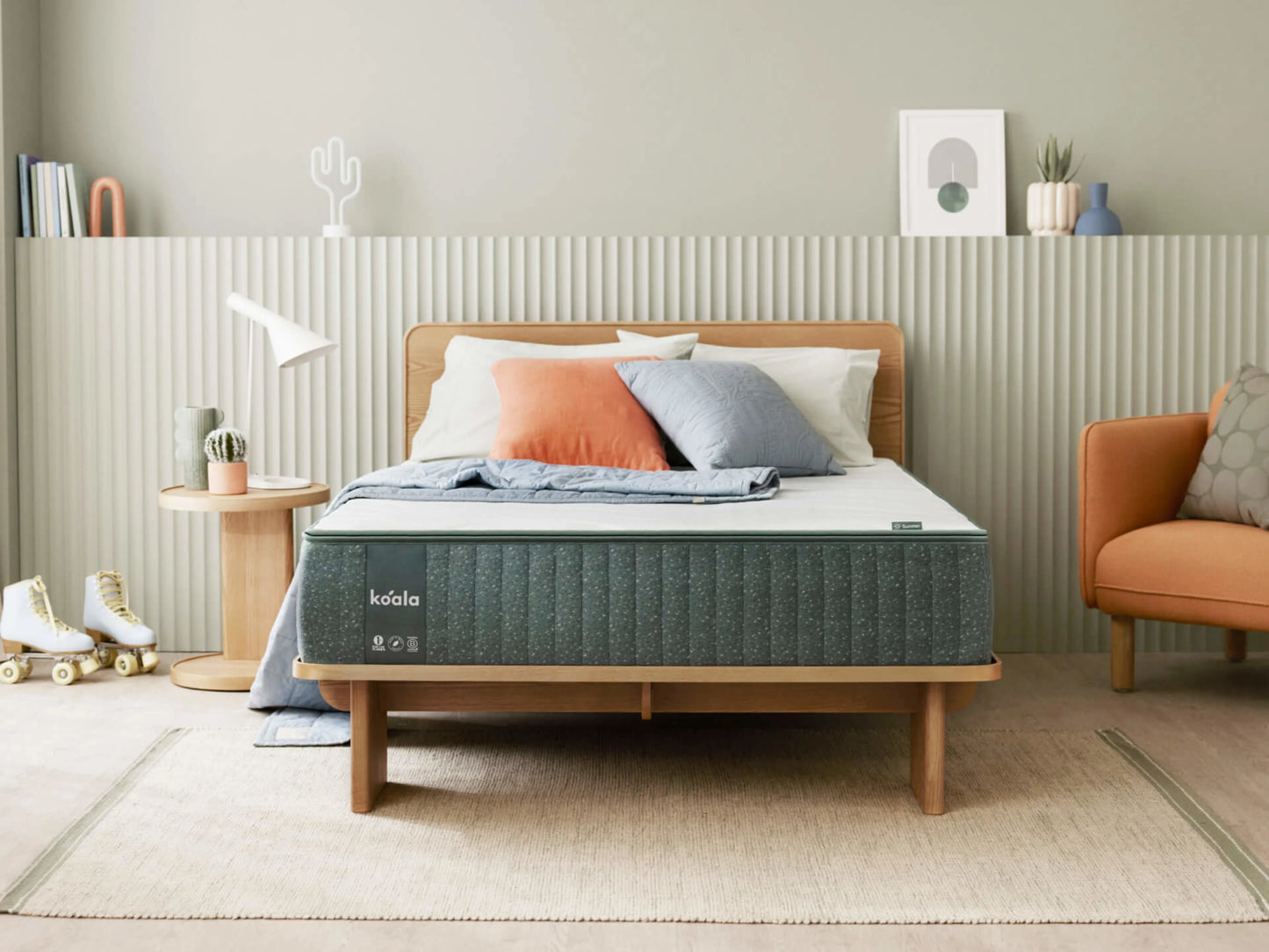 Modern bedroom with a wooden bed frame, green mattress, and mixed cushions. Orange armchair, round bedside table, and decor on light green walls.