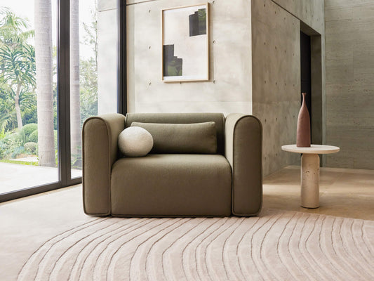 Olive-green armchair with round cushions on a beige rug. A small round table with a vase stands beside it against a concrete wall.