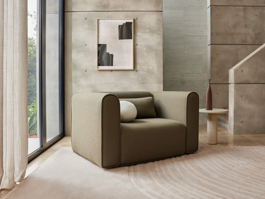 Modern living room with a light brown armchair, white cushions, and a small round table. Concrete walls, large window, and abstract art add style.