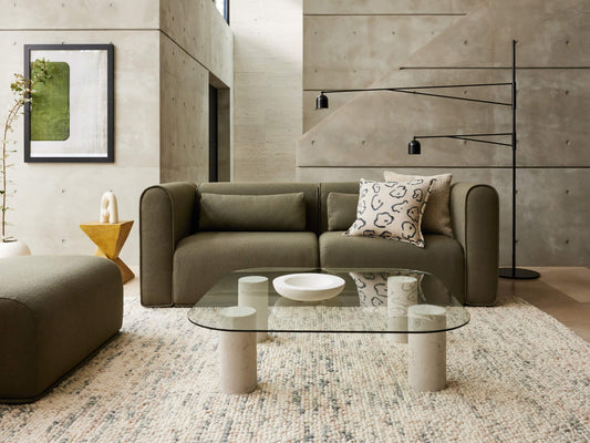 Modern living room with olive green sofa, abstract cushion, glass coffee table, and textured rug. Concrete walls and black floor lamp.