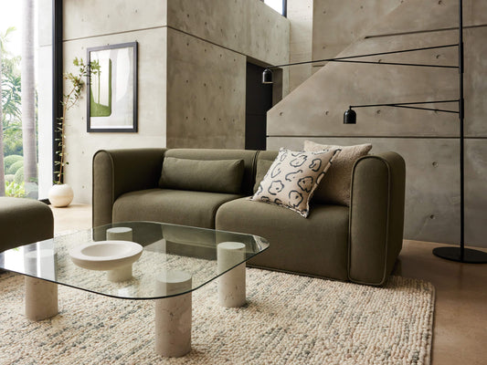 Olive green sofa with decorative cushions, glass coffee table with white cylindrical legs on a beige rug, set in a modern concrete room.