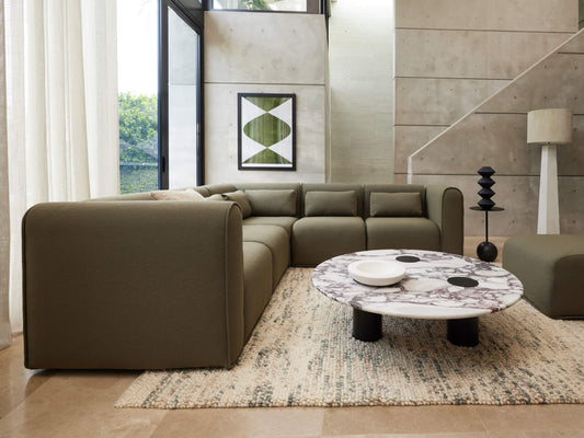 Modern living room with an olive green sectional sofa, round marble coffee table, and abstract art. Neutral rug, concrete walls, and large window.