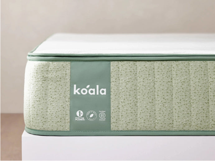 Green mattress with textured sides on a white base, featuring the "koala" logo and eco-certification icons. Background is a plain wall.