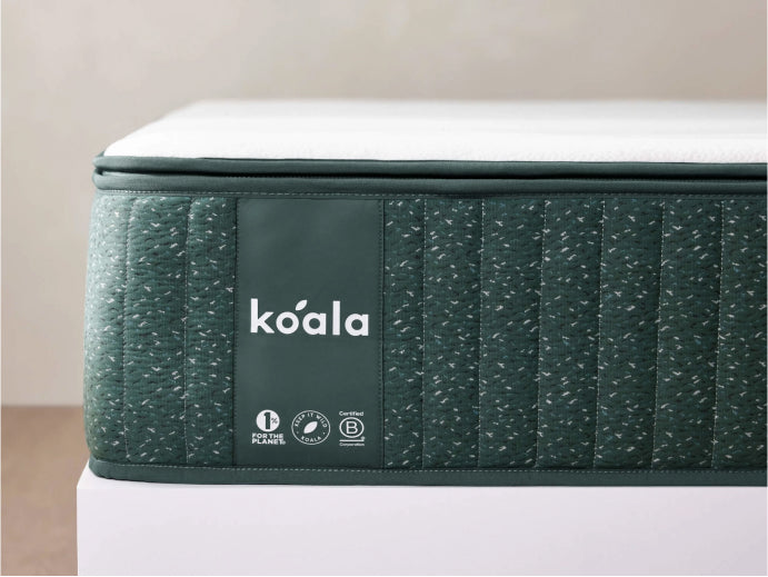 Close-up of a mattress corner with a dark green fabric cover featuring a white Koala logo and care icons.