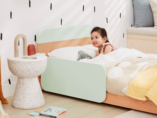 Child in a mint green toddler bed with a colourful quilt, smiling. Speckled round side table holds decor. Black dash-patterned wallpaper.