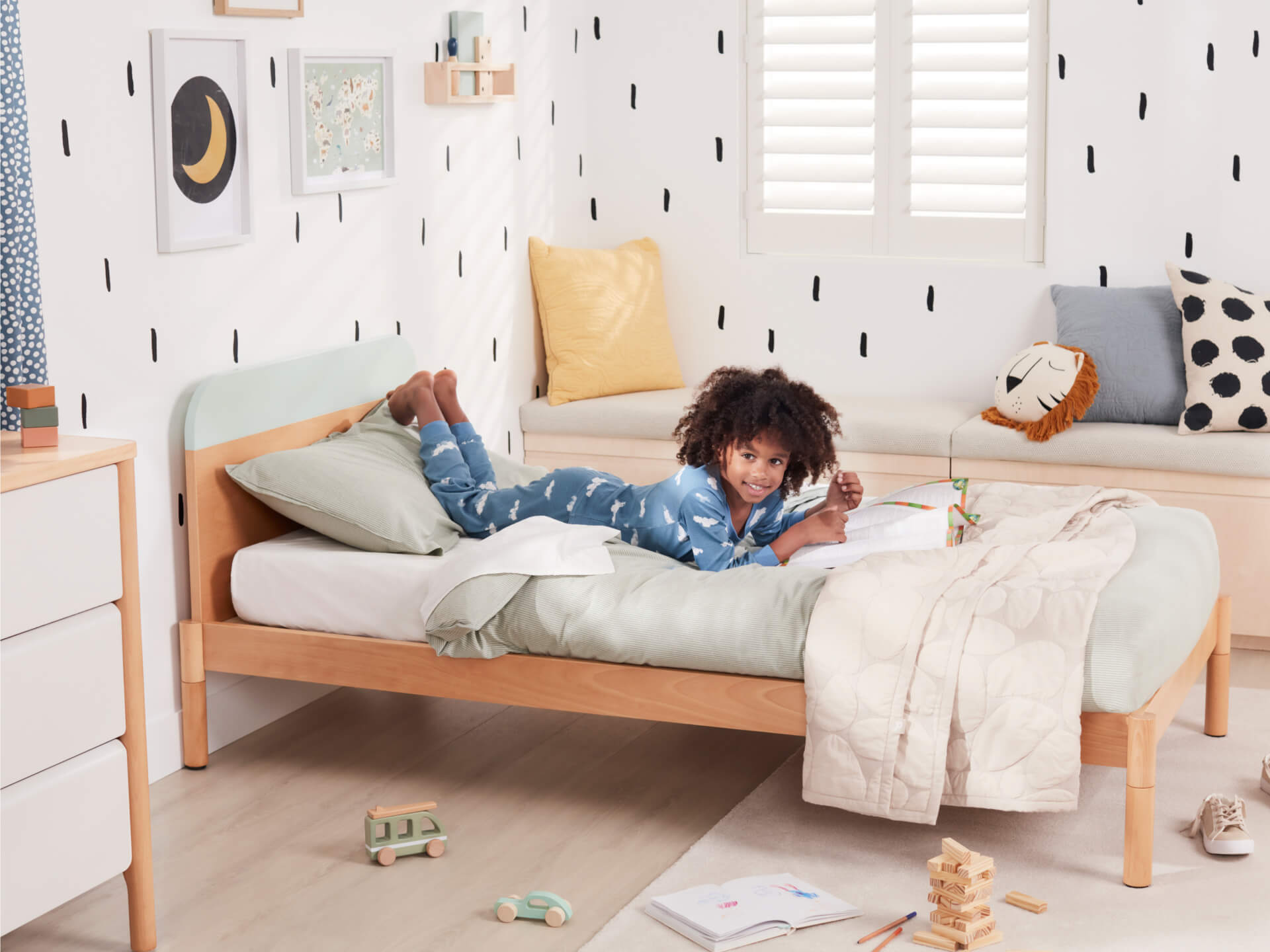 Child lying on a light wood bed with mint and white bedding, in a modern kids' room. Features playful decor, toys, and patterned wall accents.