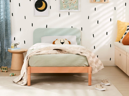 Child's bedroom with light green headboard, wooden bed frame, beige quilt, and patterned pillow. White walls with black dash accents.