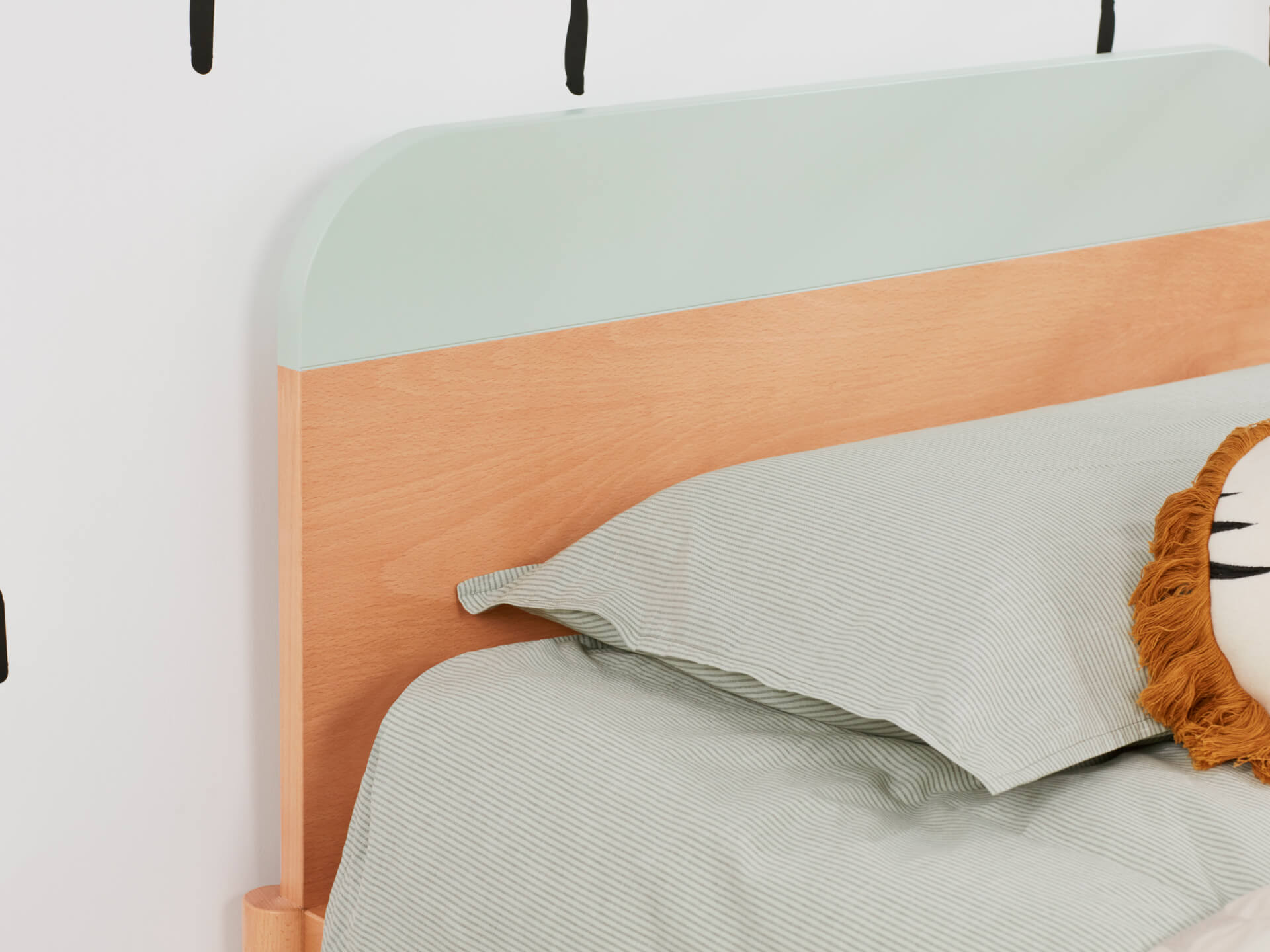 Light wood bed with pale green headboard, light green striped bedding, and a playful lion-themed pillow against a white wall.