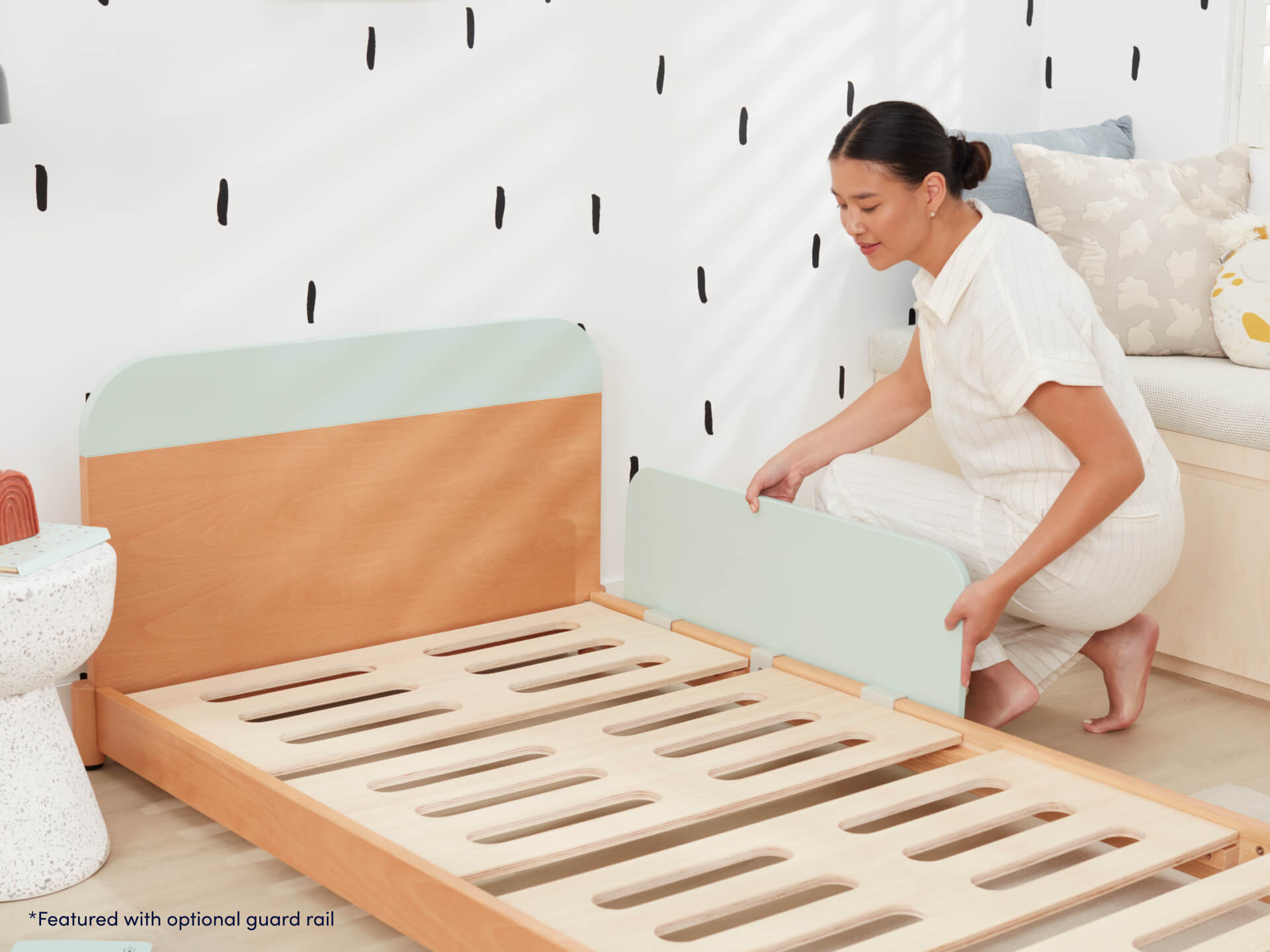 Woman assembling a light wood bed frame with mint green guard rail in a modern room with black and white wall pattern.