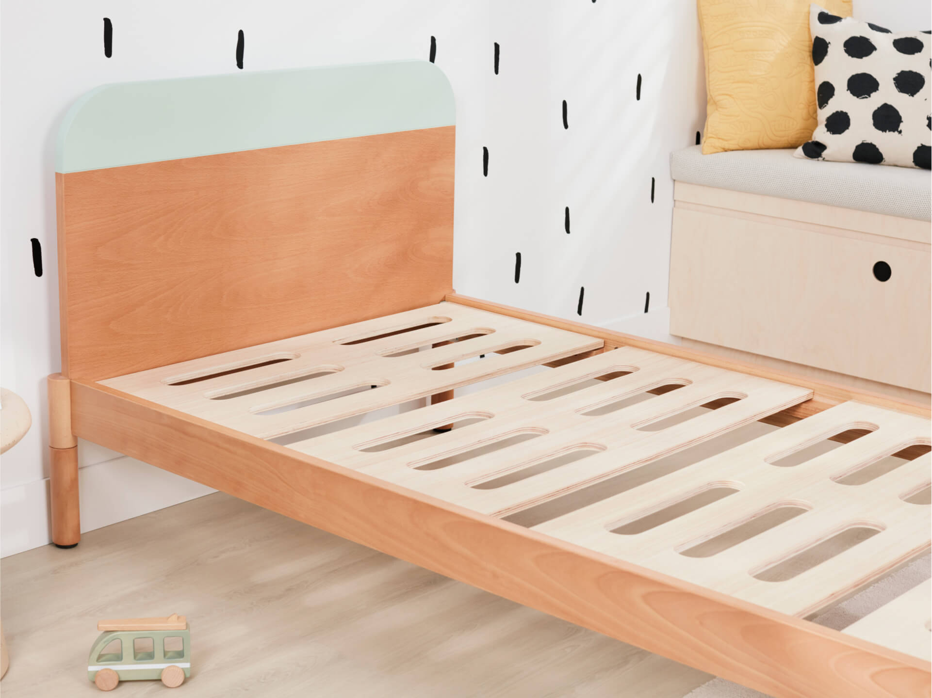 Wooden kids bed frame with mint green and natural wood headboard, slatted base. Polka dot cushion on a beige bench in the background.