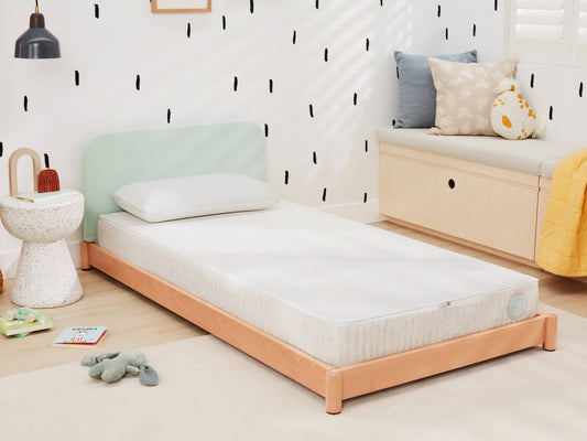 Single bed with a light green headboard and white mattress. Surrounding decor includes a side table, books, a plush toy, and a window seat.