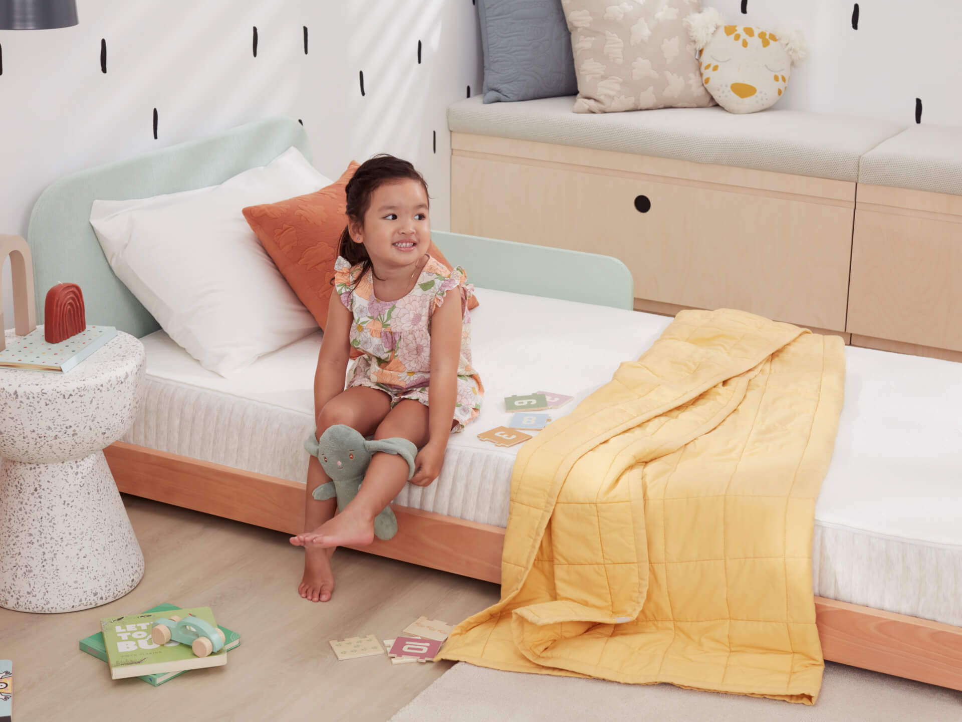 Child sitting on a light wood bed with a white mattress and yellow blanket. Side table with books, plush cushions, and playful decor.