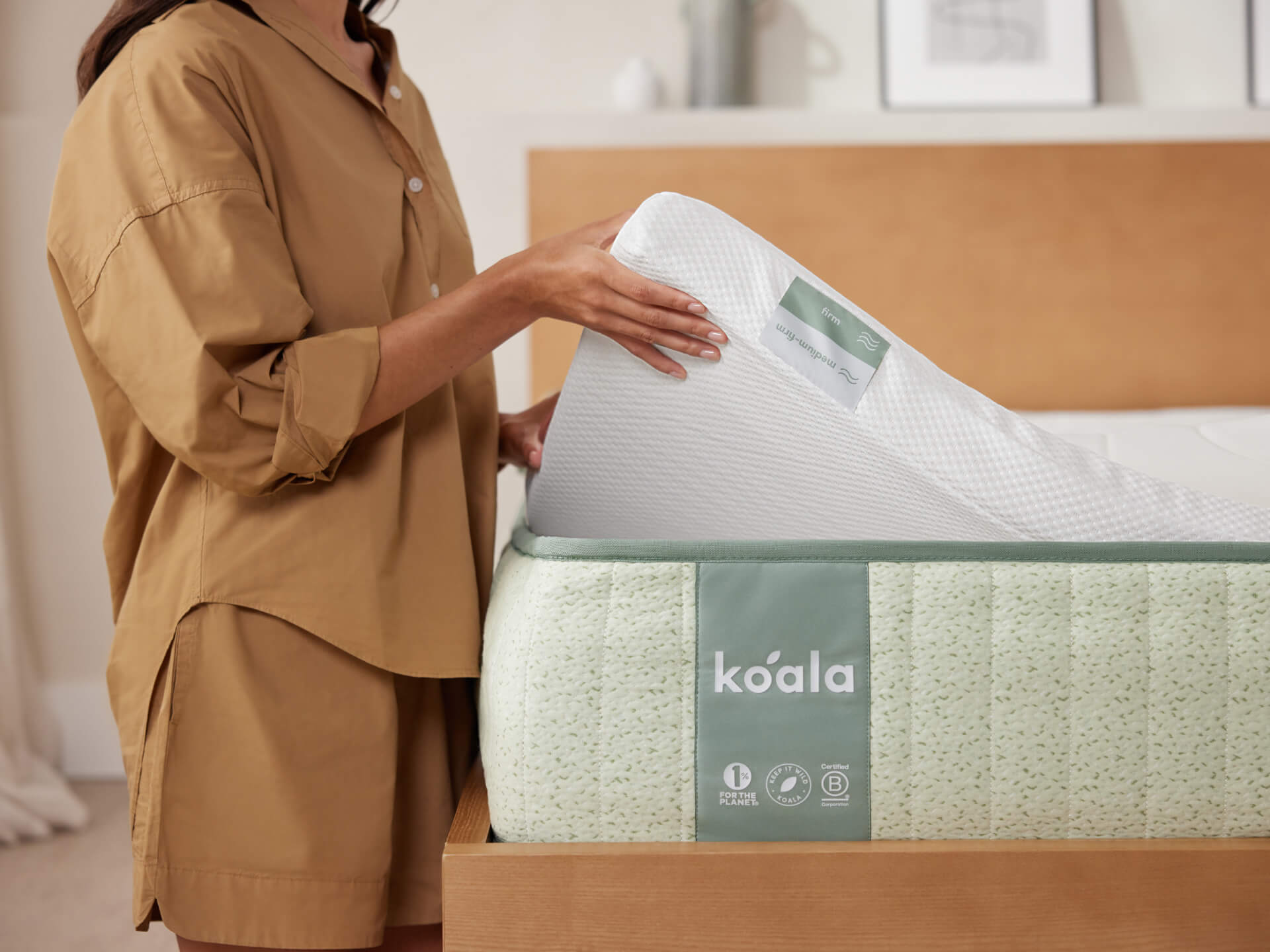 Person adjusting a white mattress topper with a mint green mattress that has a "koala" label, in a bedroom with light wood furniture.