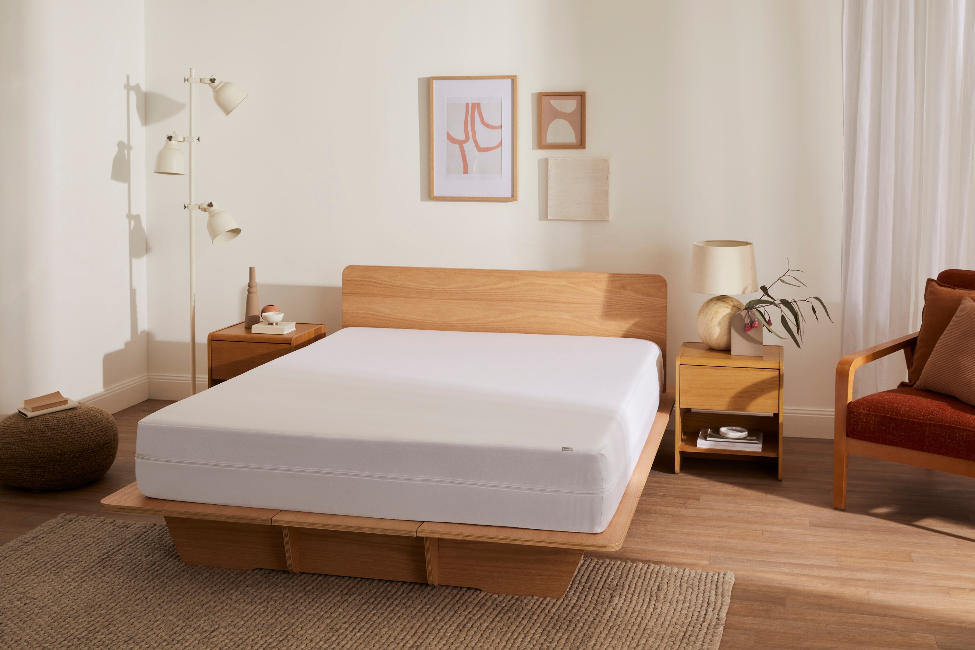 Minimalist bedroom with a light wood bed frame, white mattress, bedside tables, and a beige floor lamp. Wall art and a rust chair complete the decor.