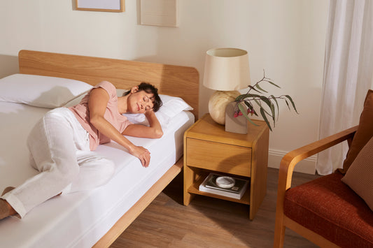 Woman sleeping on a white mattress in a minimalist bedroom with a wooden bed, bedside table with lamp, and a plant. Brown chair in the corner.