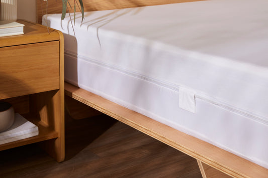 Wooden bed frame with white mattress, adjacent to a matching wooden bedside table with books and a bowl, on a wooden floor.