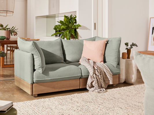 Mint green modular sofa with scattered cushions and a striped throw, sits on a light rug. Wooden floor, plants, and framed art in the background.
