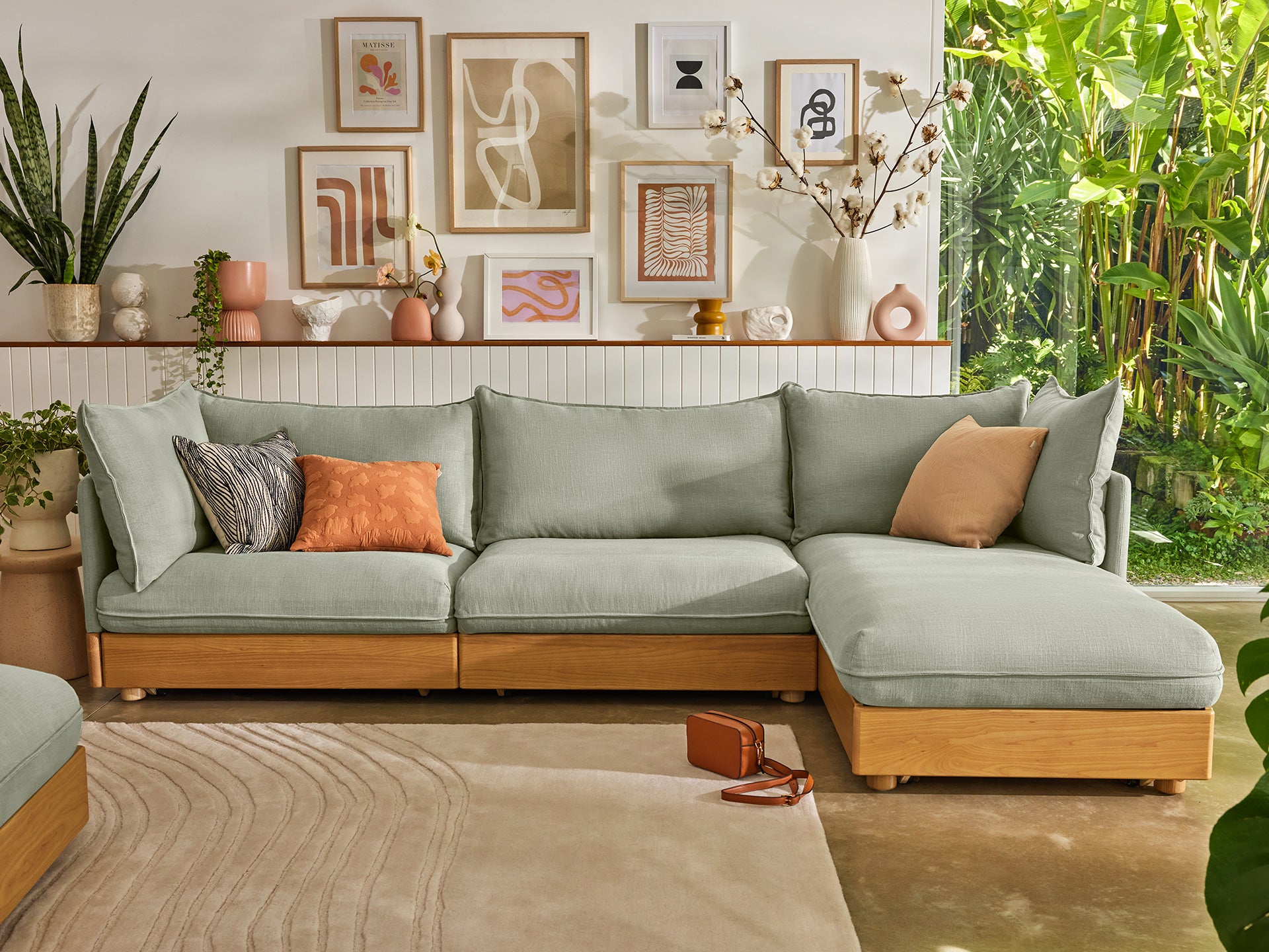 Modern living room with light green modular sofa, wooden base, decorative cushions, framed artwork, indoor plants, and sunlight streaming through.