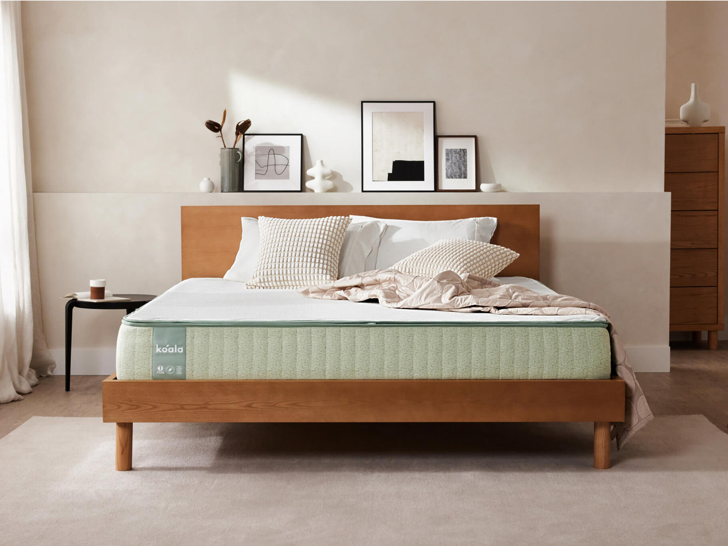 Modern bedroom with a wooden bed frame, light green mattress, and neutral bedding. Artworks and decor on white walls, and a side table.