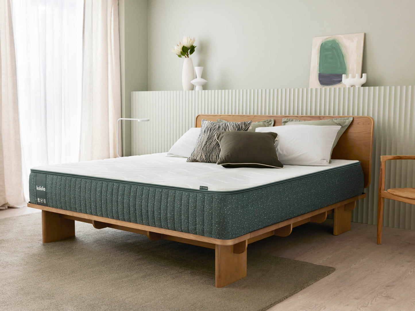 Modern bedroom with a wooden bed frame, dark green mattress, and white pillows. Light grey walls, minimalist decor, and natural light via curtains.