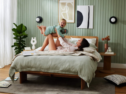 Woman and child play on a bed with light green bedding. Wooden bedside table, decor, indoor plant, and abstract art in a green-walled room.