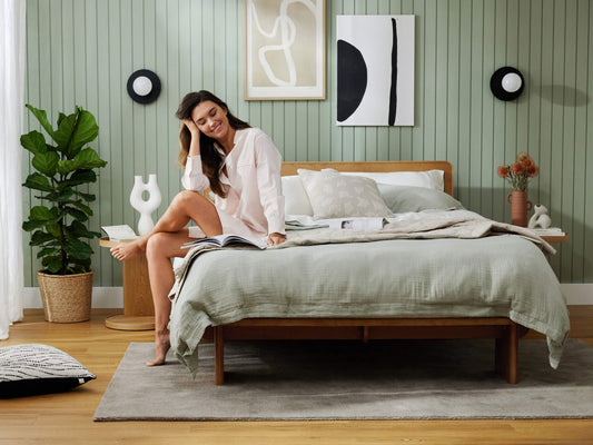 Bedroom with wooden bed, light grey bedding, woman reading, green walls, abstract art, potted plant, and circle wall decor. Casual, modern decor.