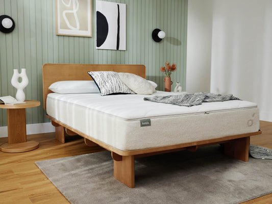 A cosy bedroom with a wooden bed frame and a white mattress. Features a green panelled wall, decorative pillows, side table, and abstract art.