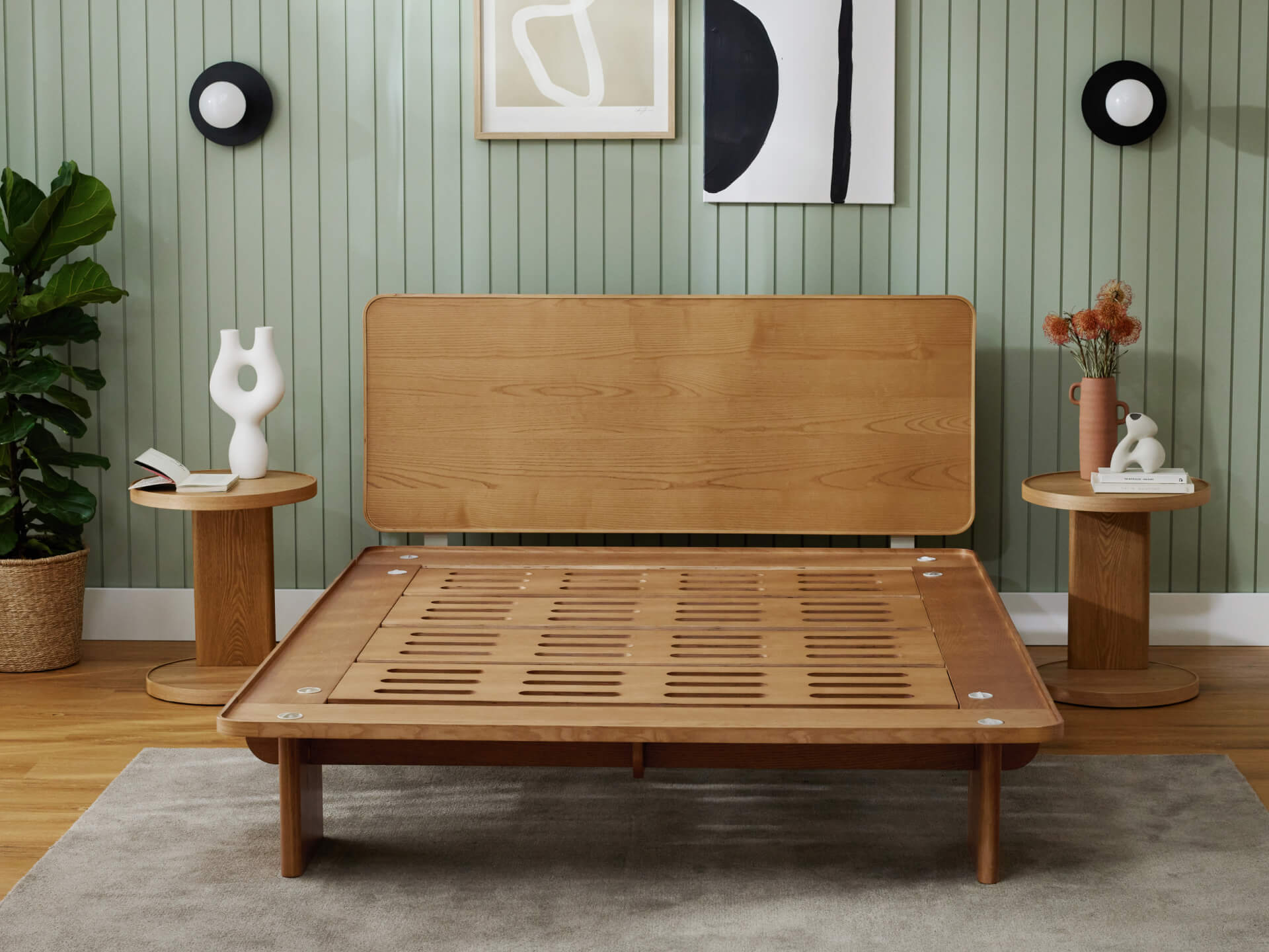 Wooden bed frame with slatted base, light brown finish, flanked by round wooden side tables. Green panelled wall, abstract art, plants.