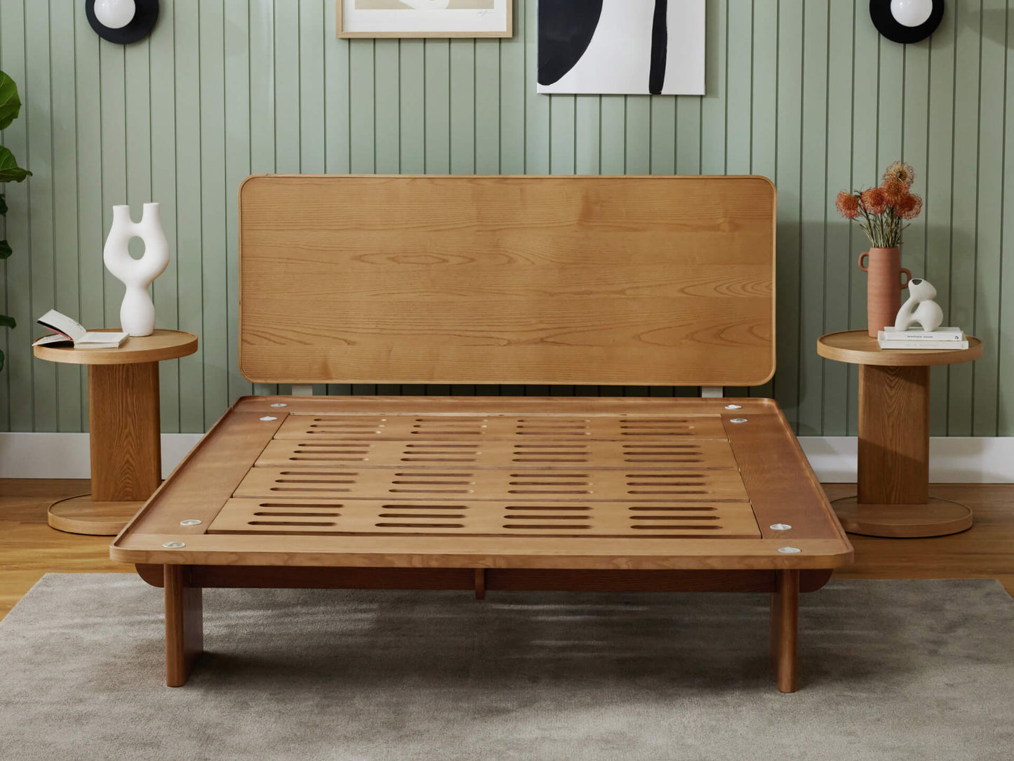Wooden bed frame with slatted base, two round wooden bedside tables, vases, green panelled wall, and light grey rug on wooden floor.