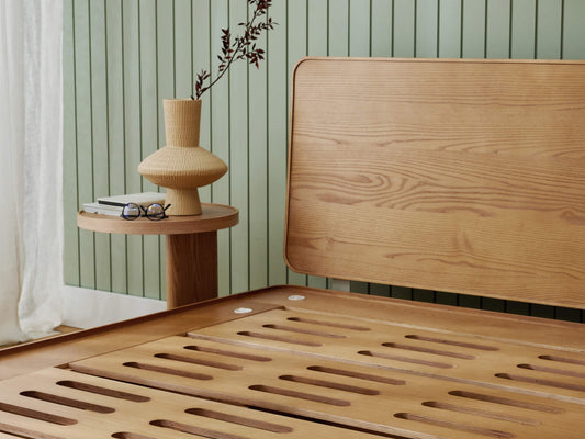 Wooden bed frame with slatted base, light wood headboard, round side table, rattan vase, and books. Background: light green panel wall.