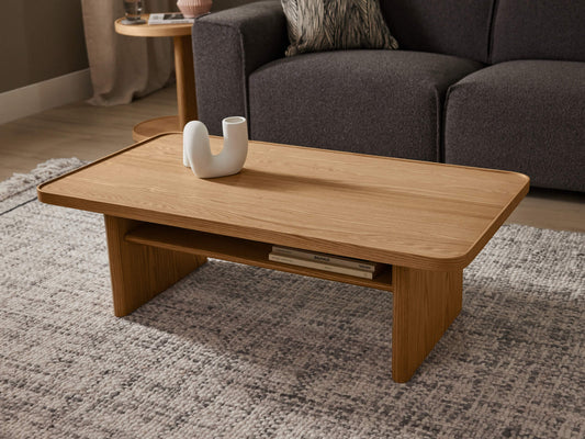 Light brown wooden coffee table on a grey rug, with a white decorative vase on top and a dark grey sofa in the background.