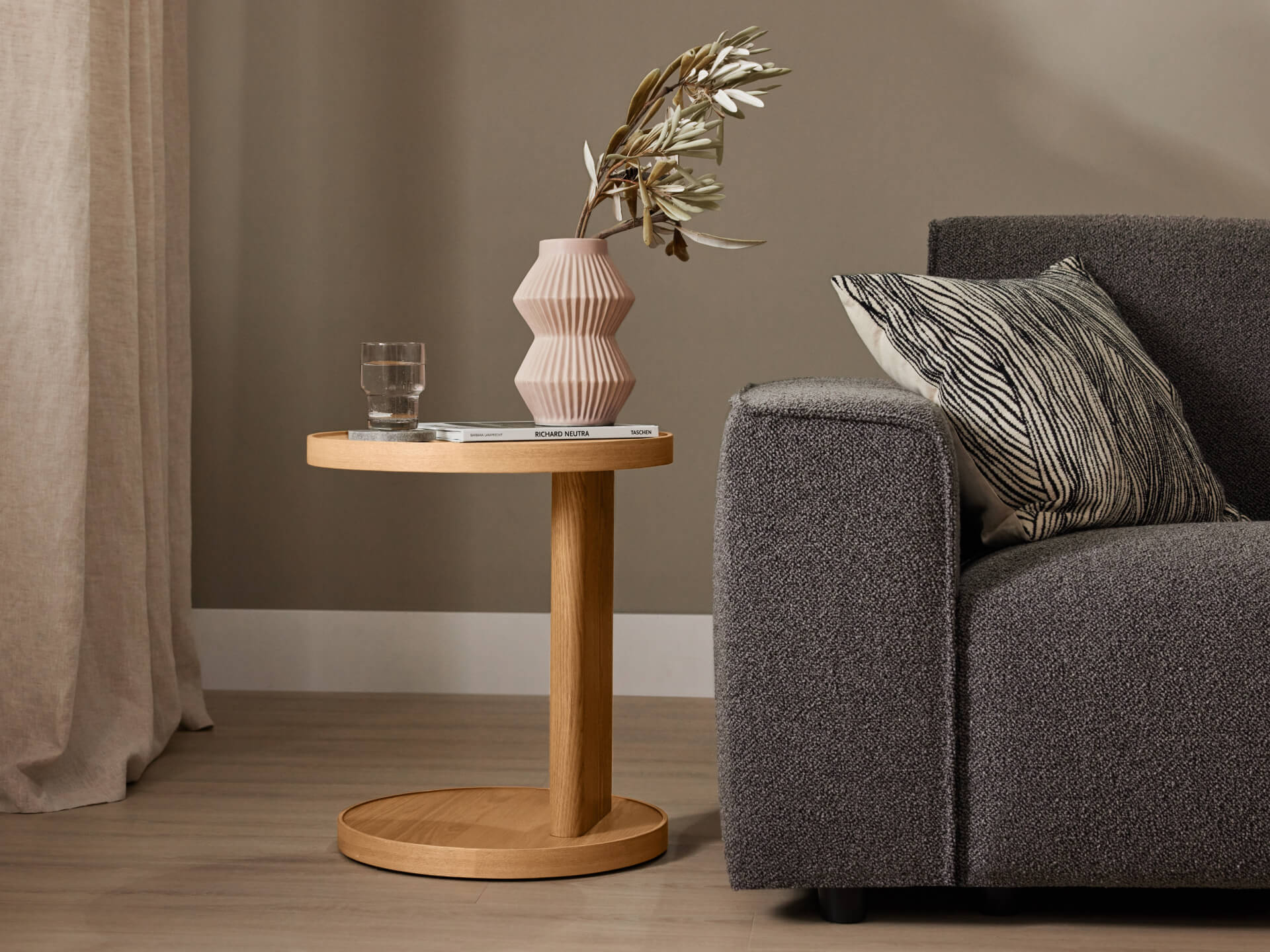 A grey couch with a patterned cushion sits beside a light wood side table holding a pink vase with foliage and a glass of water.