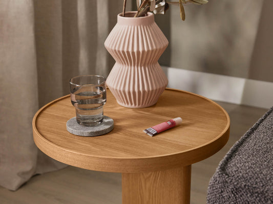Round wooden side table with a ribbed pink vase, glass of water on a coaster, and a small tube of cream. Neutral-toned fabric in the background.