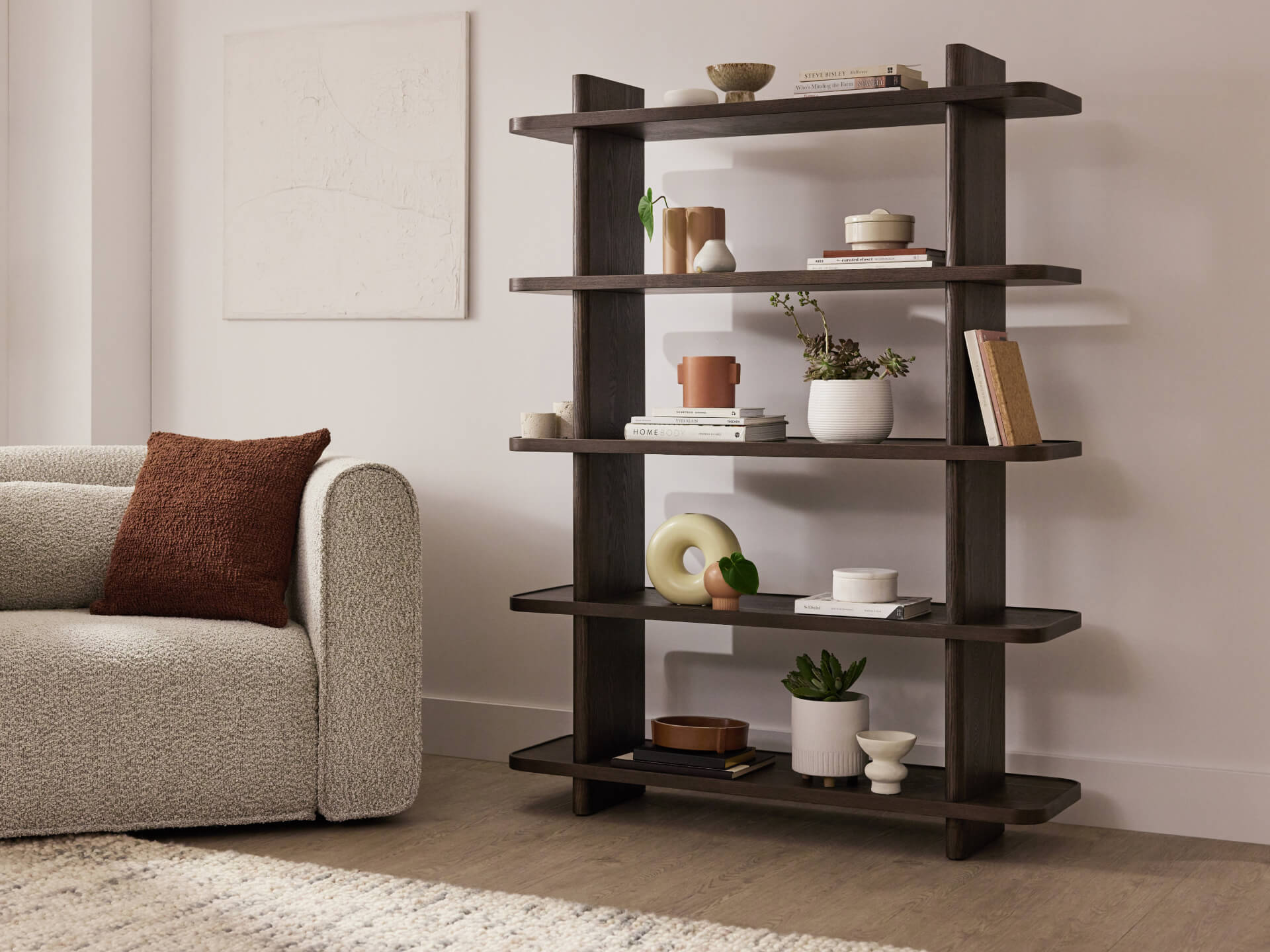 Dark brown wooden bookshelf with five shelves holds books and decor; positioned beside a textured light grey sofa with a brown cushion.