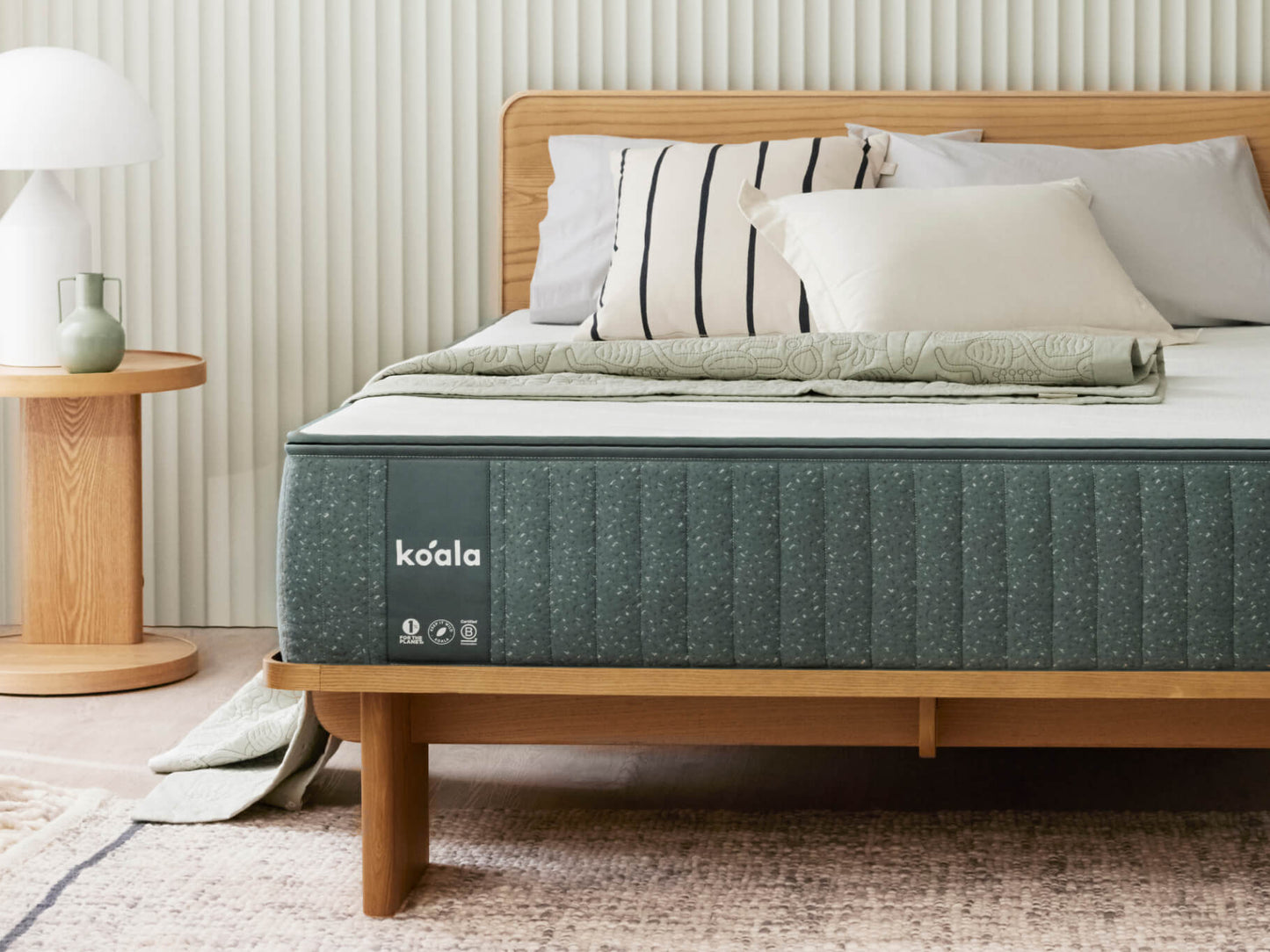 Cosy bedroom with a light wood bed frame, a dark grey mattress, and assorted cushions. A round wooden bedside table and lamp are on the left.