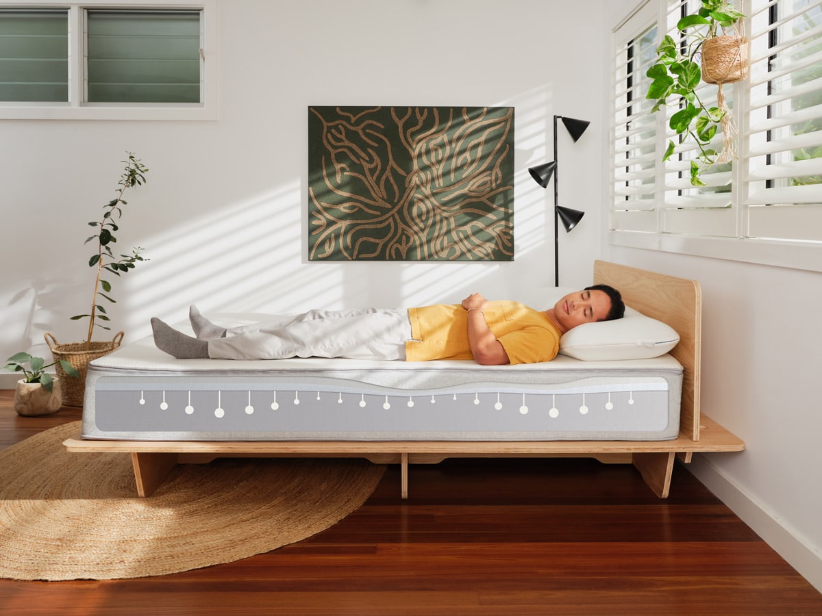 Man wearing a yellow shirt lies on a light grey mattress with a wooden base. Room features wooden floor, geometric artwork, and a hanging plant.
